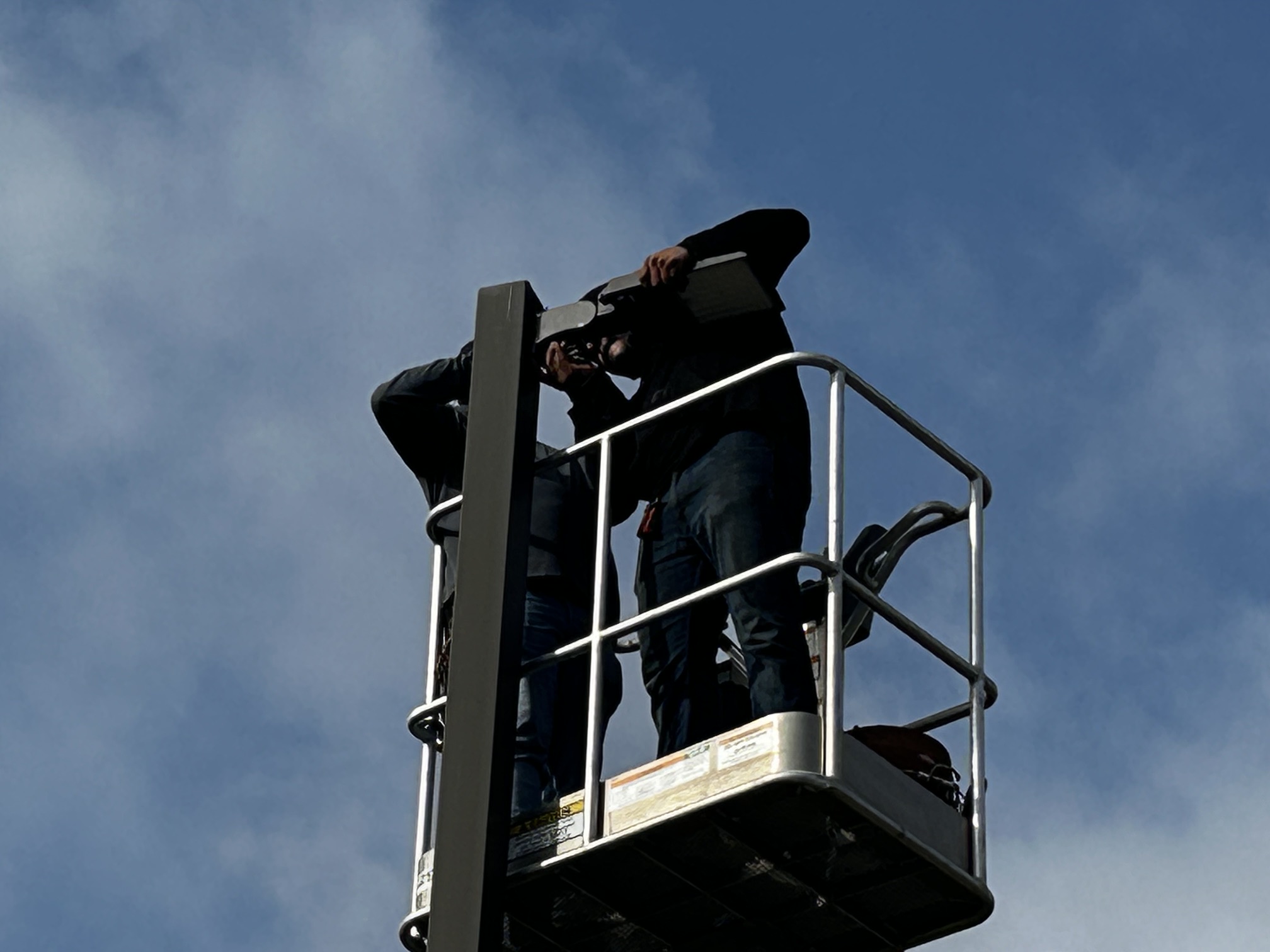 Two individuals on a lift platform, one holding a camera and the other a clipboard, against a backdrop of cloudy blue skies.
