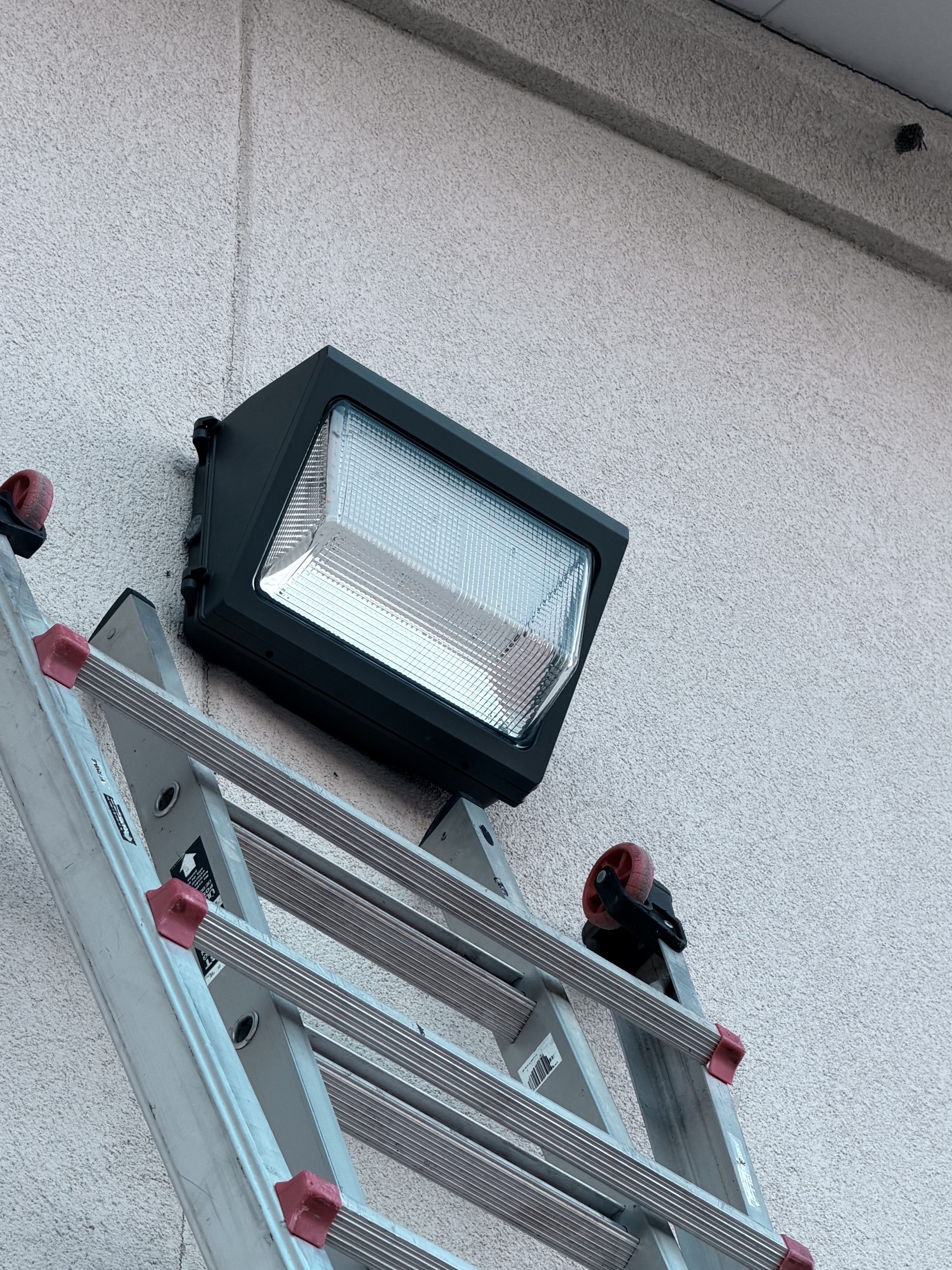 A black floodlight mounted on a textured white wall above a metal ladder.